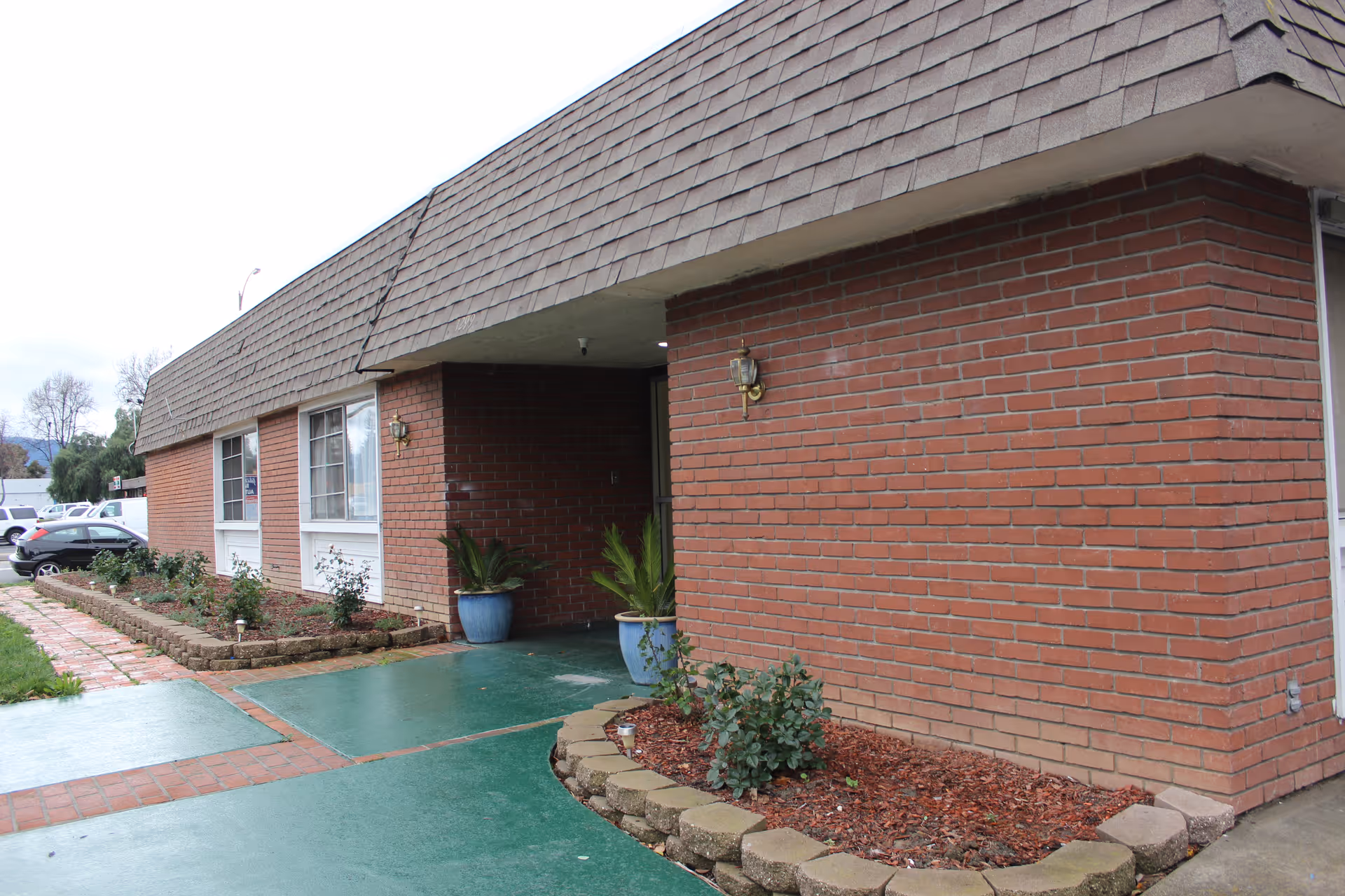 Exterior view of a single-story brick building with a shingled roof. There are two large windows and a covered entrance with two potted plants. A green walkway and a small landscaped area with plants and mulch are visible in front of the building. Several parked cars can be seen in the background.