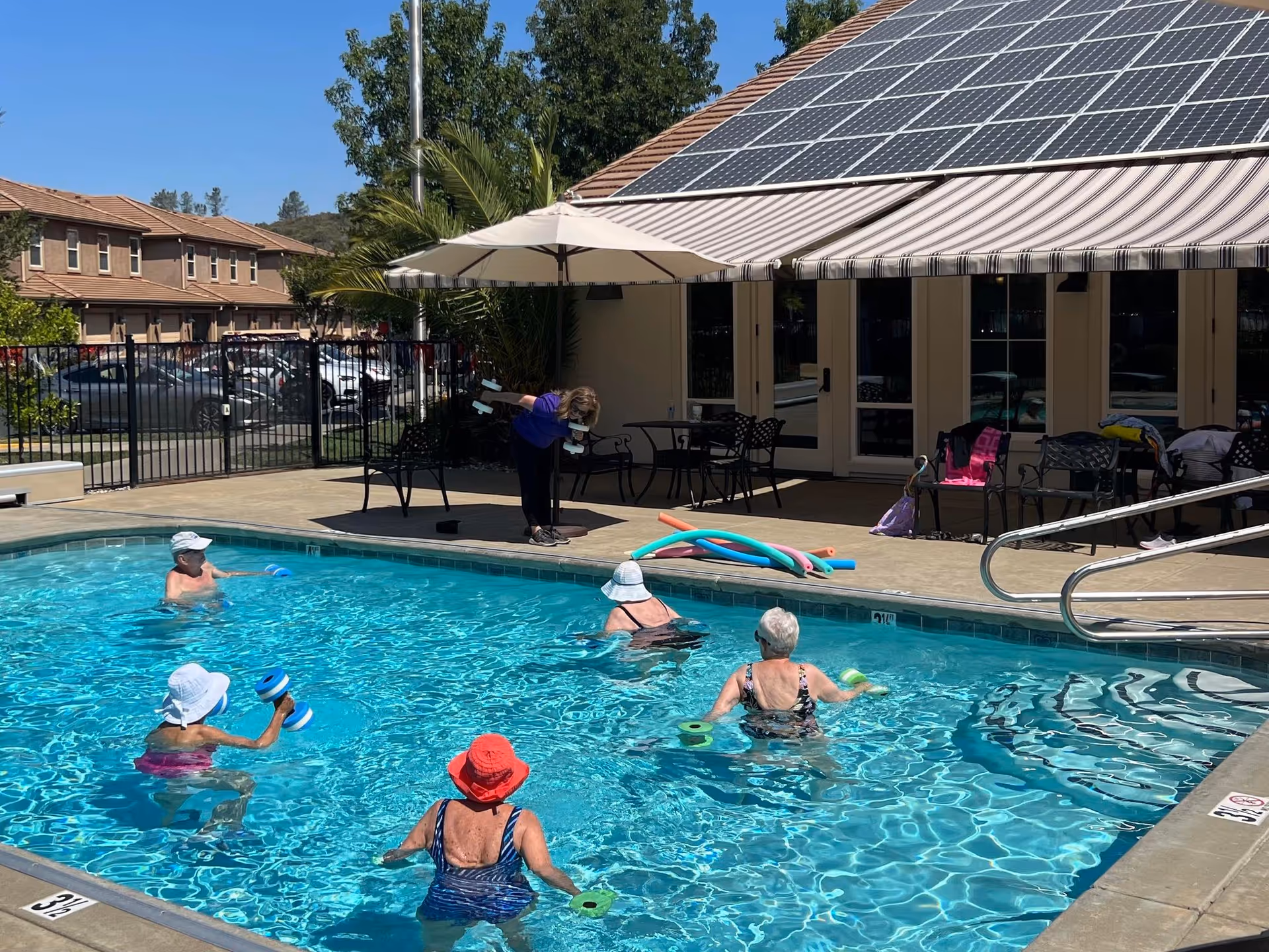 A group of elderly people participating in a water exercise class in an outdoor swimming pool at a senior living facility. An instructor stands poolside demonstrating exercises with dumbbells. The pool area is surrounded by a fence, patio furniture, and a building with solar panels on the roof.