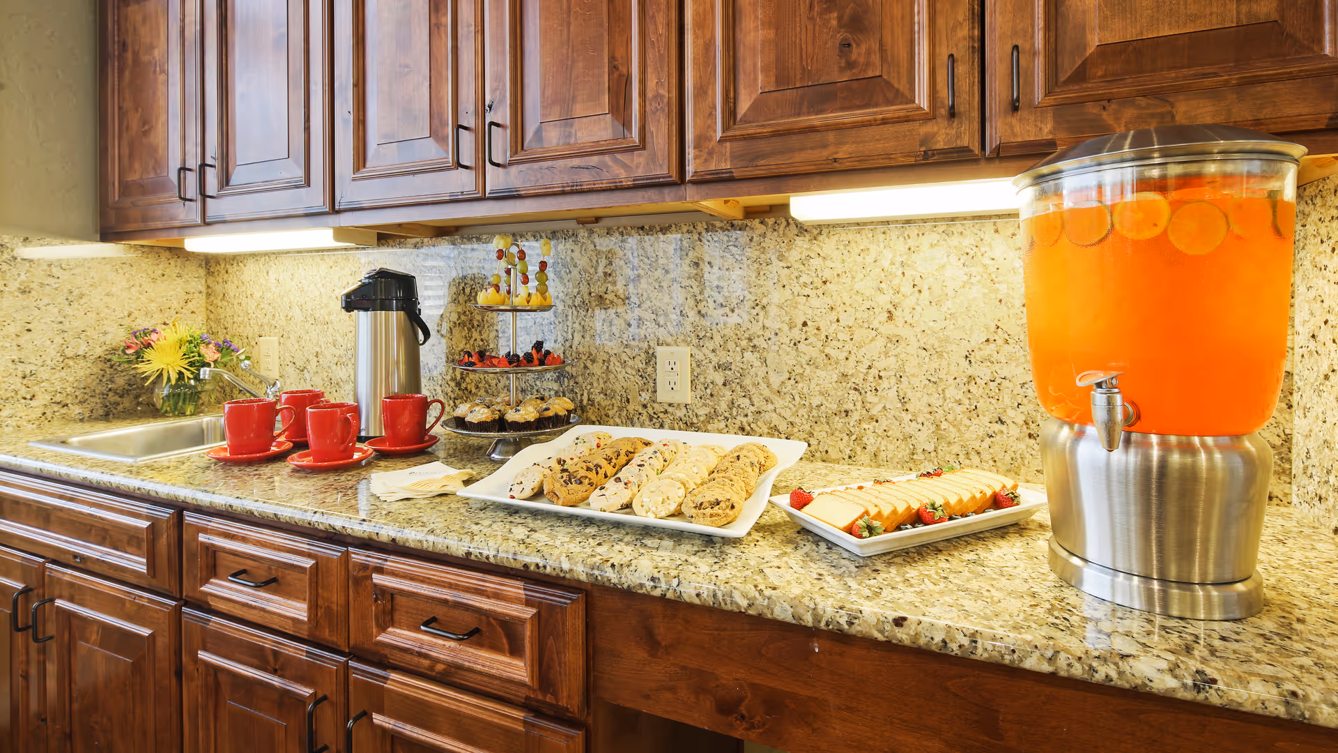 A kitchen countertop with wooden cabinets above and below. On the counter, there is a stainless steel sink, a coffee dispenser with red cups and saucers, a tiered tray with assorted pastries and fruit skewers, a plate of cookies, a plate of sliced cheese with strawberries, and a large beverage dispenser filled with orange drink and slices of citrus.