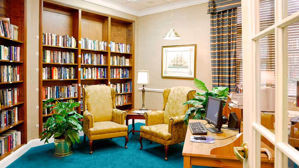 A cozy reading room with two yellow upholstered armchairs facing each other near a small wooden table with a lamp. Behind the chairs is a large wooden bookshelf filled with books. A green potted plant is placed on the floor next to the chairs. To the right, there is a wooden desk with a computer monitor, keyboard, and mouse. The room has a teal carpet, beige walls, a framed picture of a ship, and striped curtains on the window.