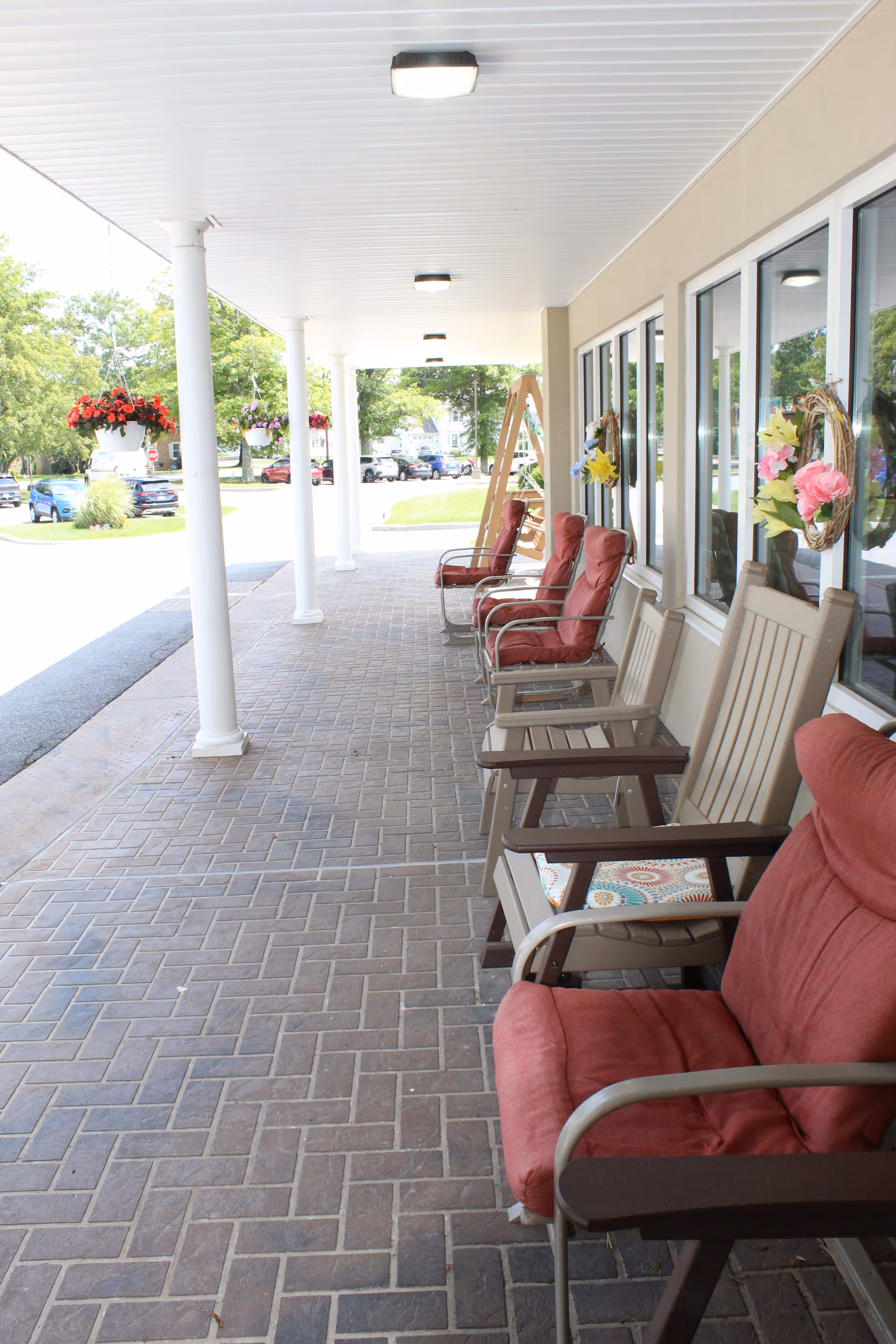 Covered outdoor patio area with cushioned chairs and hanging flower pots, adjacent to a building with windows decorated with floral wreaths. Several cars and trees are visible in the background.