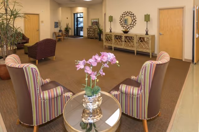 A spacious lobby seating area with two striped armchairs around a small round table holding a pink orchid, a console table and plants against the far wall.