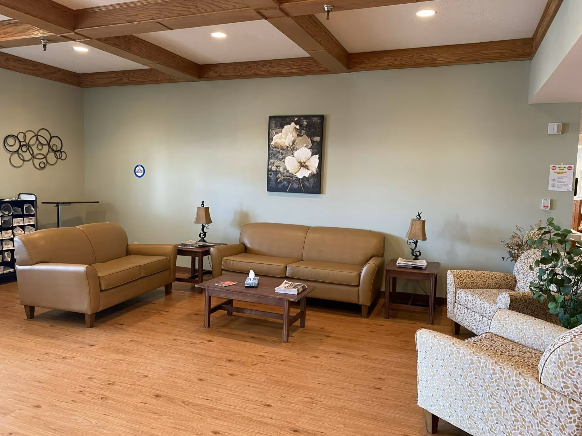 A nursing home common area with two tan leather sofas, two patterned armchairs, wooden coffee and side tables with lamps, a floral painting on the wall, and a decorative metal wall art piece. The room has wooden flooring and a ceiling with wooden beams and recessed lighting.