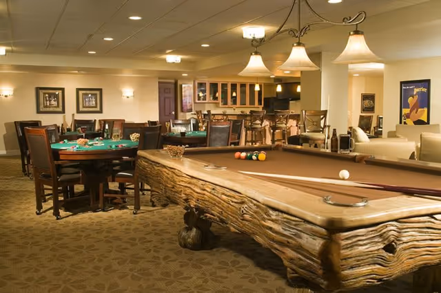 A spacious recreational room featuring a rustic wooden pool table with billiard balls and cues, several card tables with chairs, a bar area with stools, and comfortable seating. The room is warmly lit with ceiling lights and pendant lamps, and decorated with framed pictures and posters on the walls.
