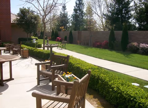 Outdoor patio area with wooden chairs and tables, surrounded by green grass, bushes, trees, and a concrete walkway. There is a brick building partially visible on the left side.