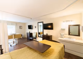 Well-lit senior care bedroom featuring a bed with an overbed table, wall-mounted TV, dresser, sink and a window with sheer curtains.