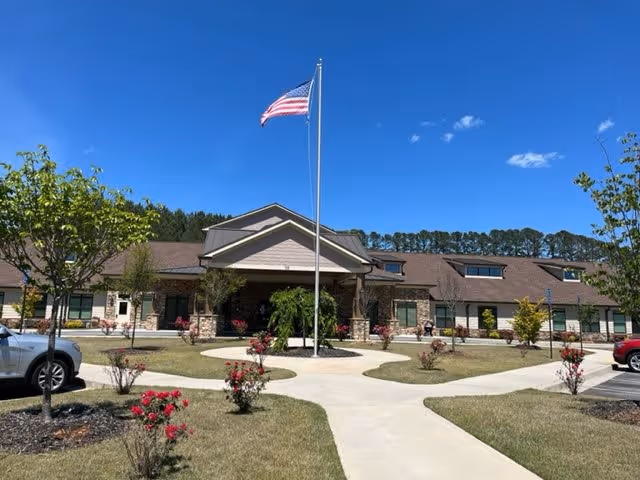 Exterior view of Dahlonega Assisted Living & Memory Care facility with a clear blue sky, an American flag on a flagpole in front, landscaped garden with small trees and red flowers, and a paved walkway leading to the entrance.