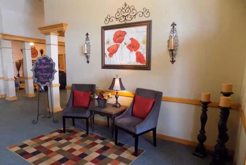 A cozy seating area in a senior living facility with two gray chairs featuring red cushions, a small wooden table with a lamp and a flower arrangement, a colorful checkered rug, and wall decorations including a framed painting of red poppies and two candle sconces. A decorative sign on a stand reads 'Welcome to Mackenzie Place'.