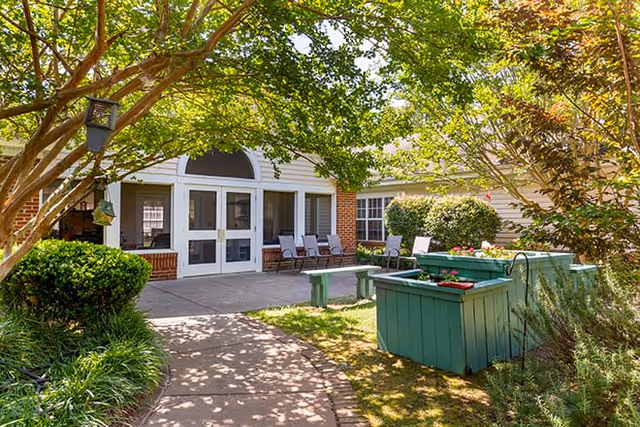 Outdoor patio area at Brookdale Winston-Salem with a paved walkway, green planter boxes, several chairs arranged near the building entrance, and trees providing shade.