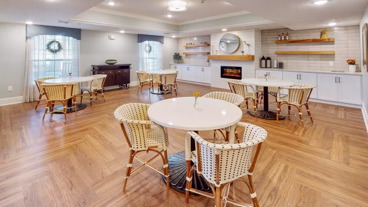 A bright and spacious dining area with several round white tables surrounded by wicker chairs with wooden legs. The room features wood-patterned flooring, large windows with sheer curtains, a modern electric fireplace with a round mirror above it, and white cabinetry with open wooden shelves on the walls.
