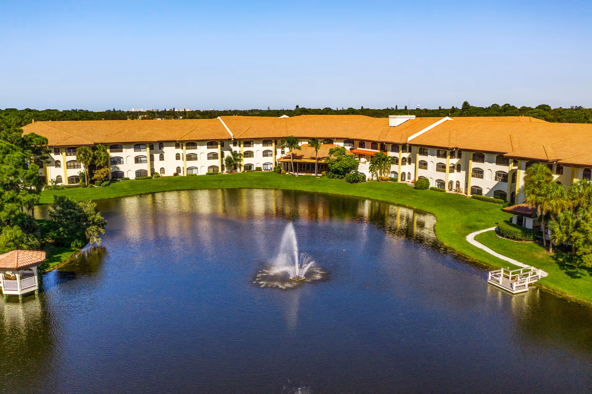 A large senior living facility building with a terracotta roof surrounding a pond with a central water fountain. The building is three stories high with multiple windows and balconies. There is a small gazebo on the left side near the pond and a small dock on the right side. The area is surrounded by green grass and trees under a clear blue sky.