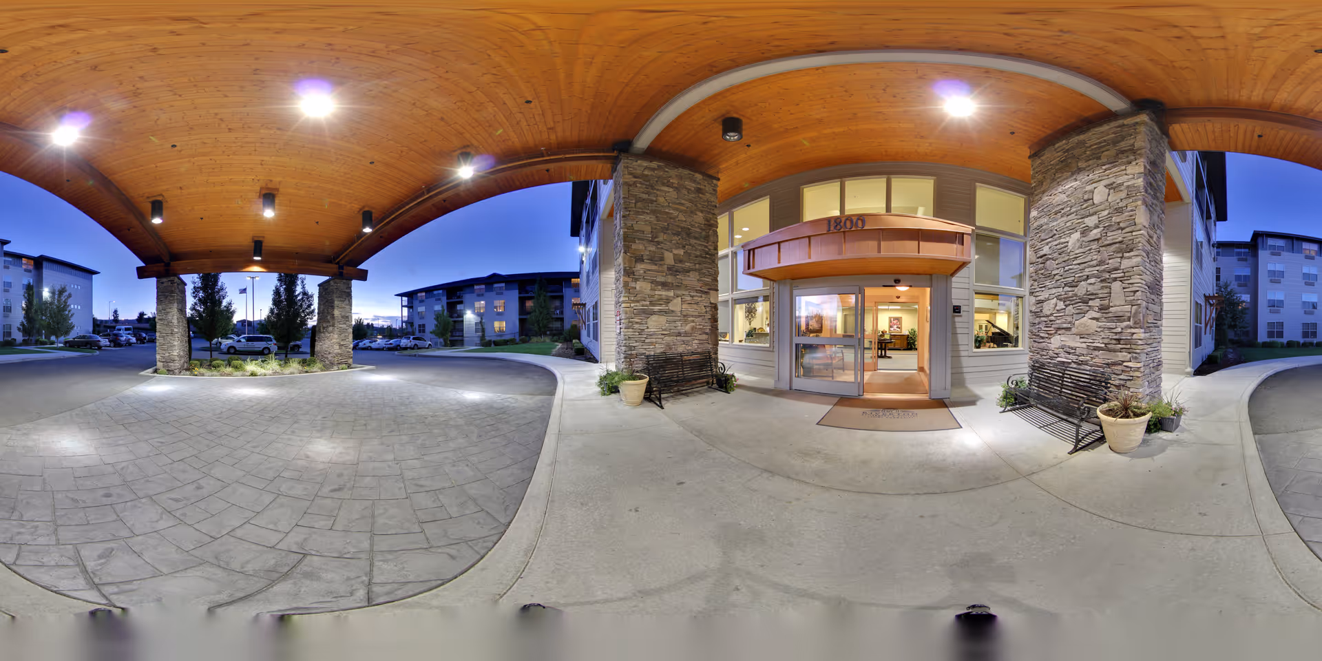 Covered entrance area of a senior living facility at dusk with stone pillars, wooden ceiling, benches, potted plants, and glass doors leading inside. Surrounding buildings and parked cars are visible in the background.