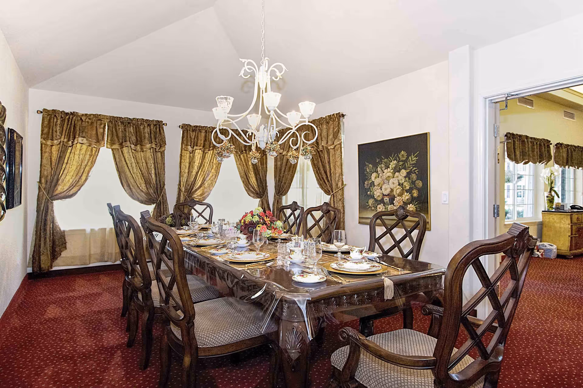 A formal dining room with a long wooden table covered with a clear protective cover, set with fine china, glassware, and a floral centerpiece. The room has red carpet flooring, ornate wooden chairs, gold patterned curtains on the windows, a white chandelier hanging above the table, and a floral painting on the wall. An open doorway leads to another room with similar decor.