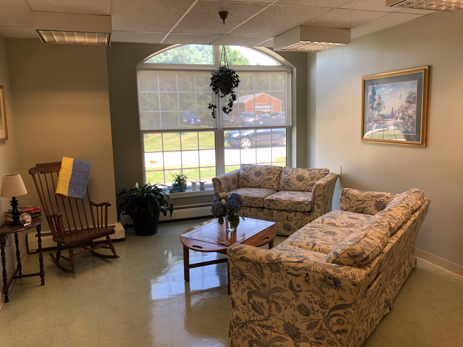 A cozy sitting area with two floral patterned sofas facing a wooden coffee table with a vase of flowers. A large window with an arched top lets in natural light. There is a wooden rocking chair with a folded quilt draped over the back, a small side table with a lamp, books, and a clock. A framed painting hangs on the wall, and a hanging plant is suspended in front of the window.