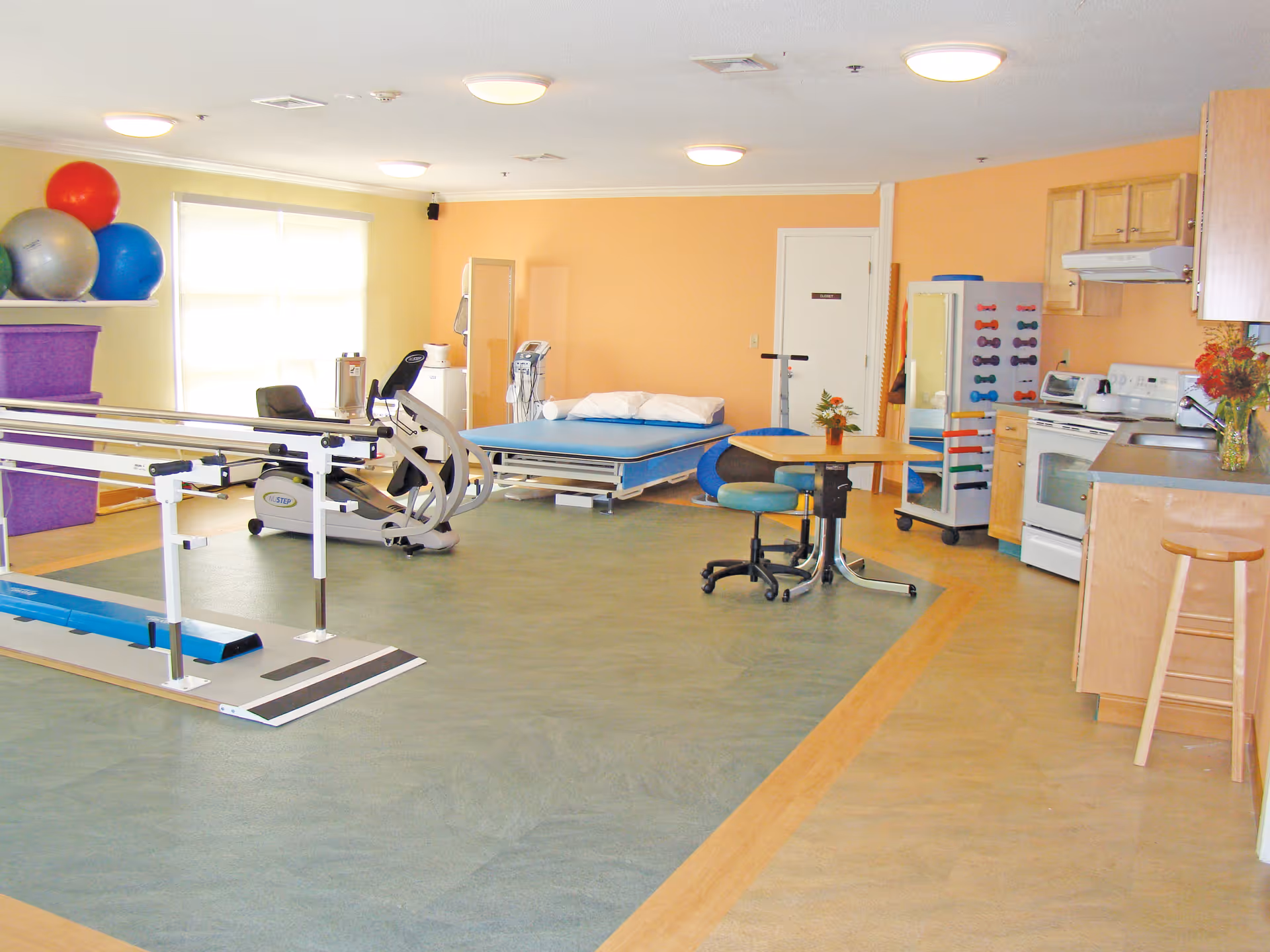 A spacious room with physical therapy equipment including parallel bars, an exercise bike, therapy balls, and a therapy bed. There is also a small table with chairs, a rack of colorful dumbbells, and a kitchenette area with a stove, microwave, and cabinets. The walls are painted in light yellow and orange tones.