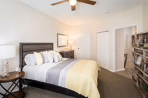 A bedroom with a neatly made bed featuring a striped comforter in shades of gray, white, and yellow. There is a wooden headboard, a round side table with a lamp, and a dresser with a lamp and mirror. The room has beige walls, a ceiling fan, carpeted floor, and two white closet doors. A wooden shelving unit with decorative items is visible on the right side.
