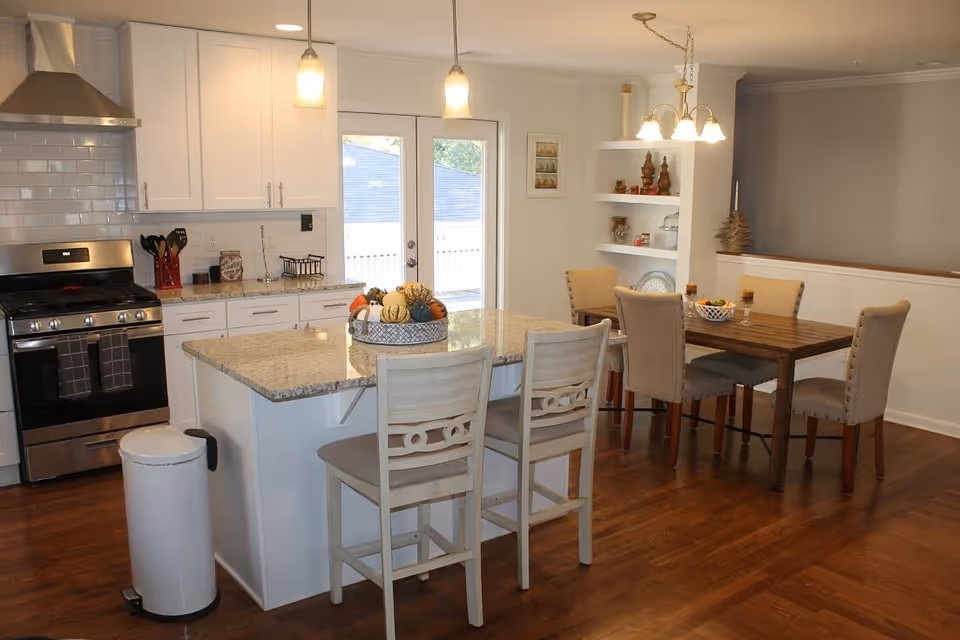 A modern kitchen and dining area featuring a stainless steel stove with a range hood, white cabinets, and a granite countertop island with two chairs. The dining area has a wooden table with four upholstered chairs and decorative shelves on the wall. Pendant lights hang above the island and dining table, and there are double glass doors leading outside.
