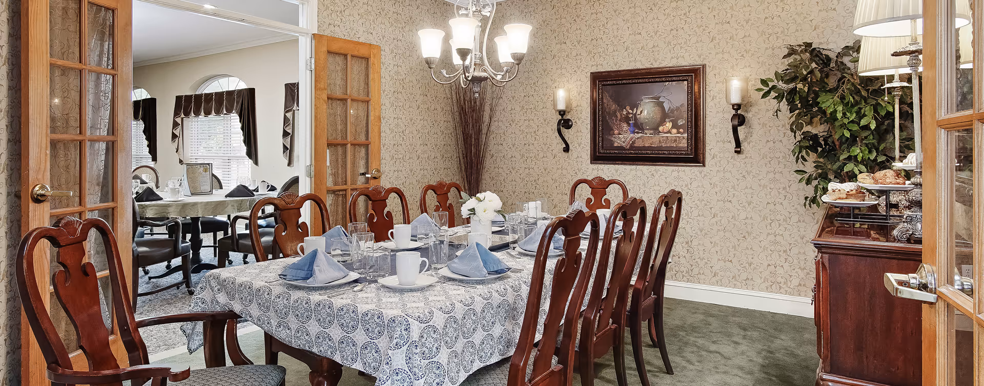A formal dining room with a rectangular table covered with a patterned tablecloth, set with plates, cups, glasses, and folded blue napkins. The room features wooden chairs with carved backs, a chandelier overhead, patterned wallpaper, a framed painting on the wall, and a sideboard with pastries and a lamp. French doors open to another dining area with round tables and windows with valances.