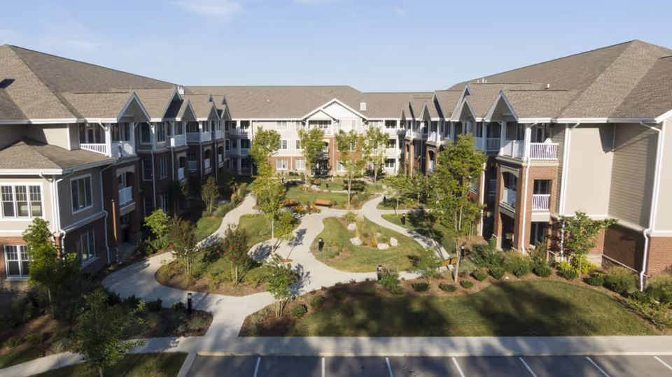 Aerial view of a senior living facility courtyard with walking paths, benches, trees, and landscaped greenery surrounded by a three-story building with balconies and windows under a clear blue sky.