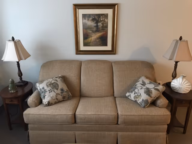 A beige upholstered sofa with two floral-patterned cushions is centered against a light-colored wall. On either side of the sofa are wooden side tables, each with a table lamp. Above the sofa hangs a framed painting depicting a tree-lined path.