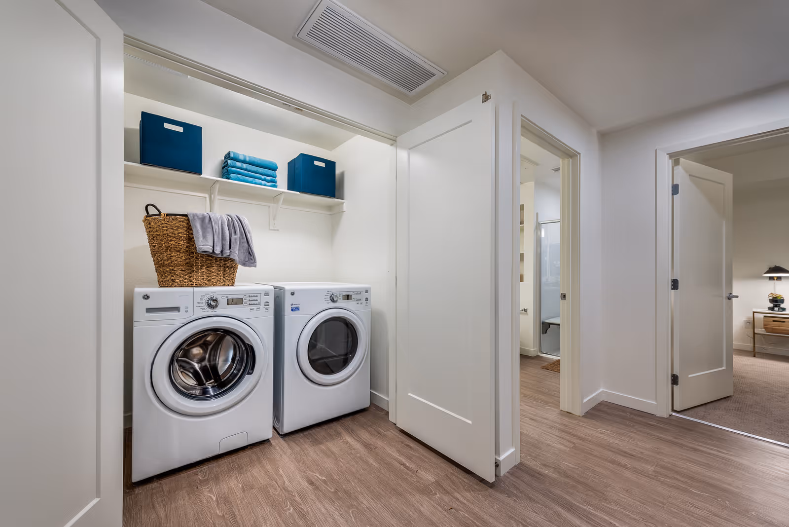 Laundry area with a front-loading washing machine and dryer side by side, a wicker basket with a gray towel on top, and a shelf above holding two blue storage bins and folded blue towels. The area is adjacent to a hallway with doors leading to other rooms.