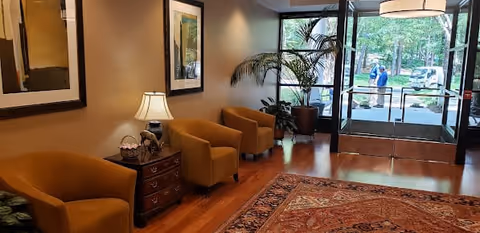 A cozy waiting area with three mustard-colored armchairs arranged along a wall with framed artwork. A wooden side table with a lamp and decorative items is placed between two chairs. A large potted plant is near the glass entrance doors, through which trees and two people outside are visible. The floor is wooden with a patterned rug covering part of it.