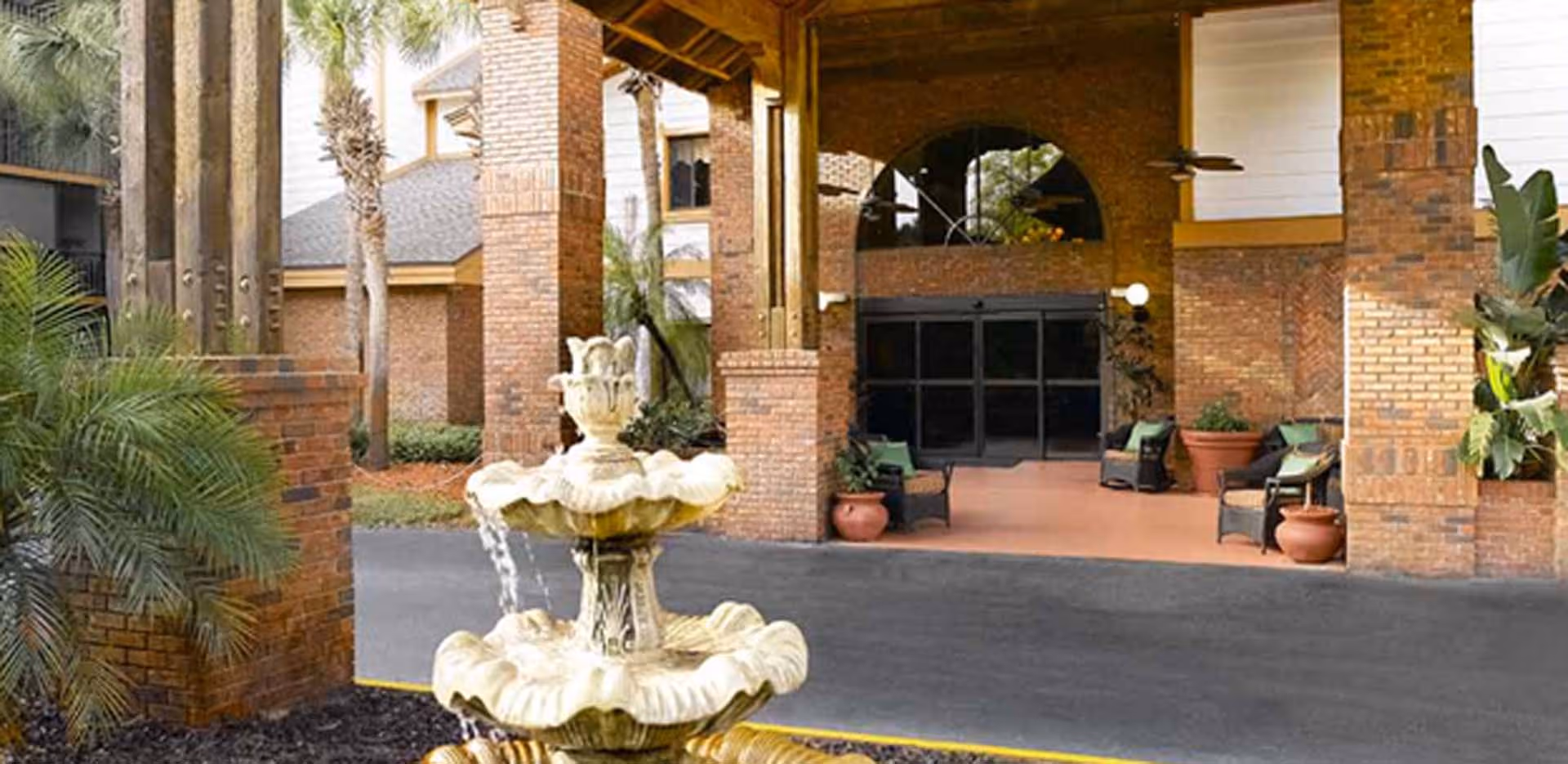 Entrance area of Vitality Living Baypoint Village featuring a covered driveway with brick pillars, a decorative two-tier water fountain in the foreground, potted plants, and outdoor seating with chairs and cushions near the entrance doors.