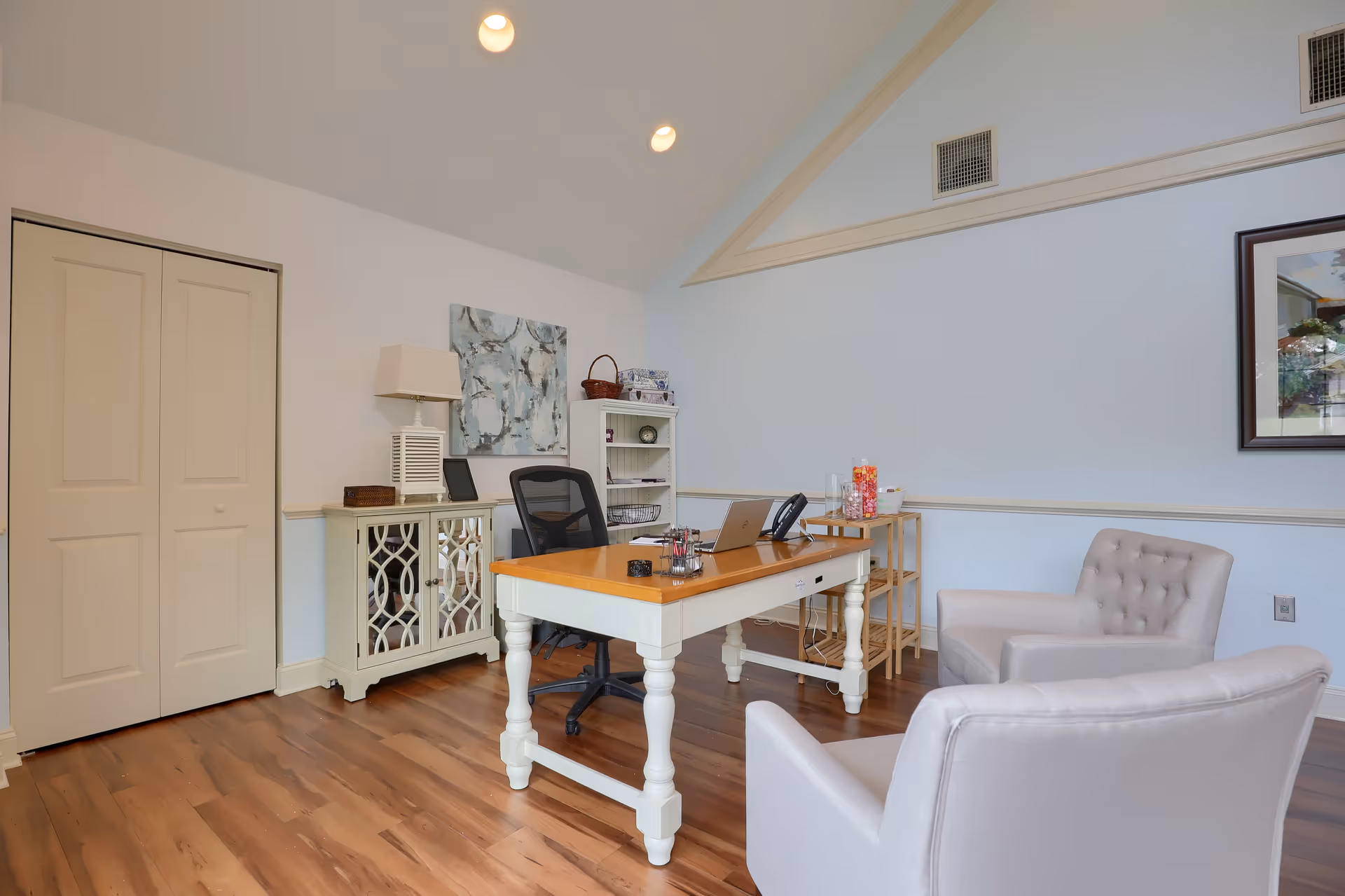 A bright office space with wooden flooring, a white desk with a wooden top, a black office chair, two light gray armchairs, a small white cabinet with a lamp and decorative items, a small shelving unit, and a framed picture on the light blue wall.