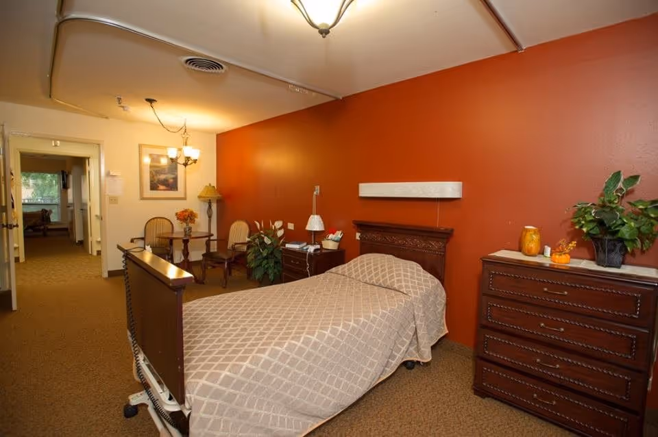 A senior living facility bedroom with a single bed covered in a beige patterned blanket, a wooden headboard and footboard, a wooden dresser with decorative items and a potted plant on top, a small table with two chairs, a floor lamp, and a red accent wall behind the bed. The room has carpeted flooring and a ceiling light fixture.