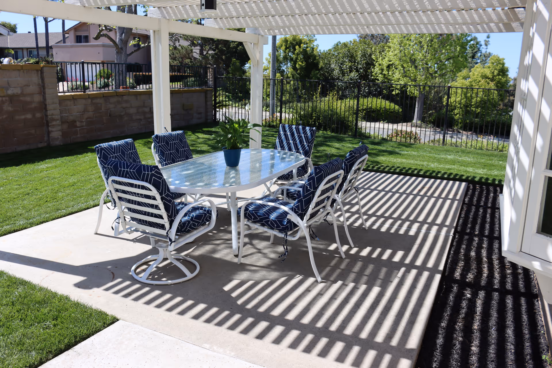 Outdoor patio area with a white pergola casting striped shadows over a glass-top table surrounded by six white metal chairs with blue patterned cushions. The patio is adjacent to a grassy lawn and enclosed by a black metal fence with trees and shrubs in the background.