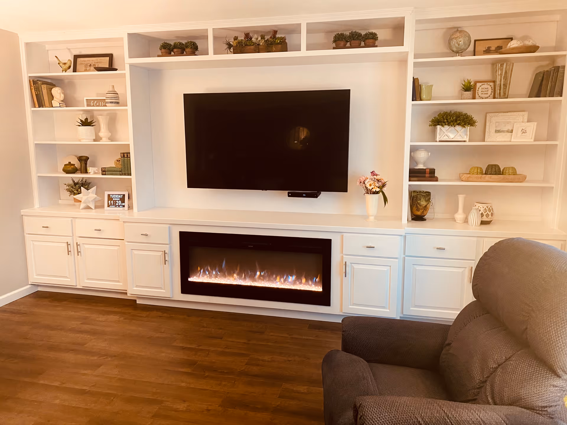 A cozy living room area featuring a large flat-screen TV mounted above a modern electric fireplace. Surrounding the TV and fireplace are white built-in shelves and cabinets decorated with various plants, books, and decorative items. A comfortable gray recliner chair is positioned in the foreground on a wooden floor.