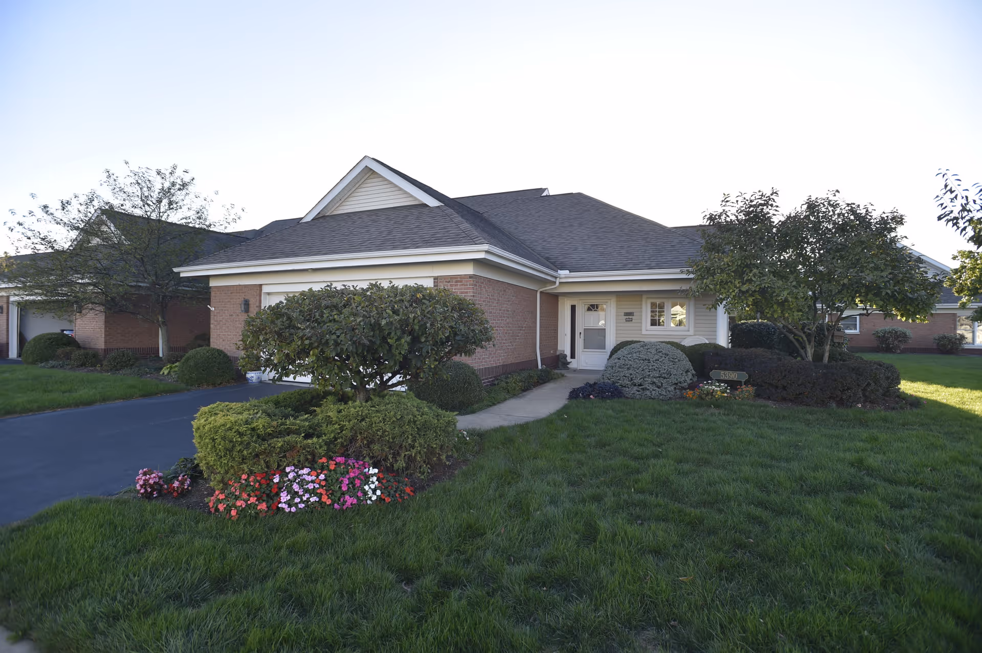 Exterior view of a single-story brick and siding building with a gable roof, surrounded by well-maintained green grass, bushes, and colorful flowers. A paved driveway and a concrete walkway lead to a white front door with windows on either side. Trees and additional buildings are visible in the background under a clear sky.