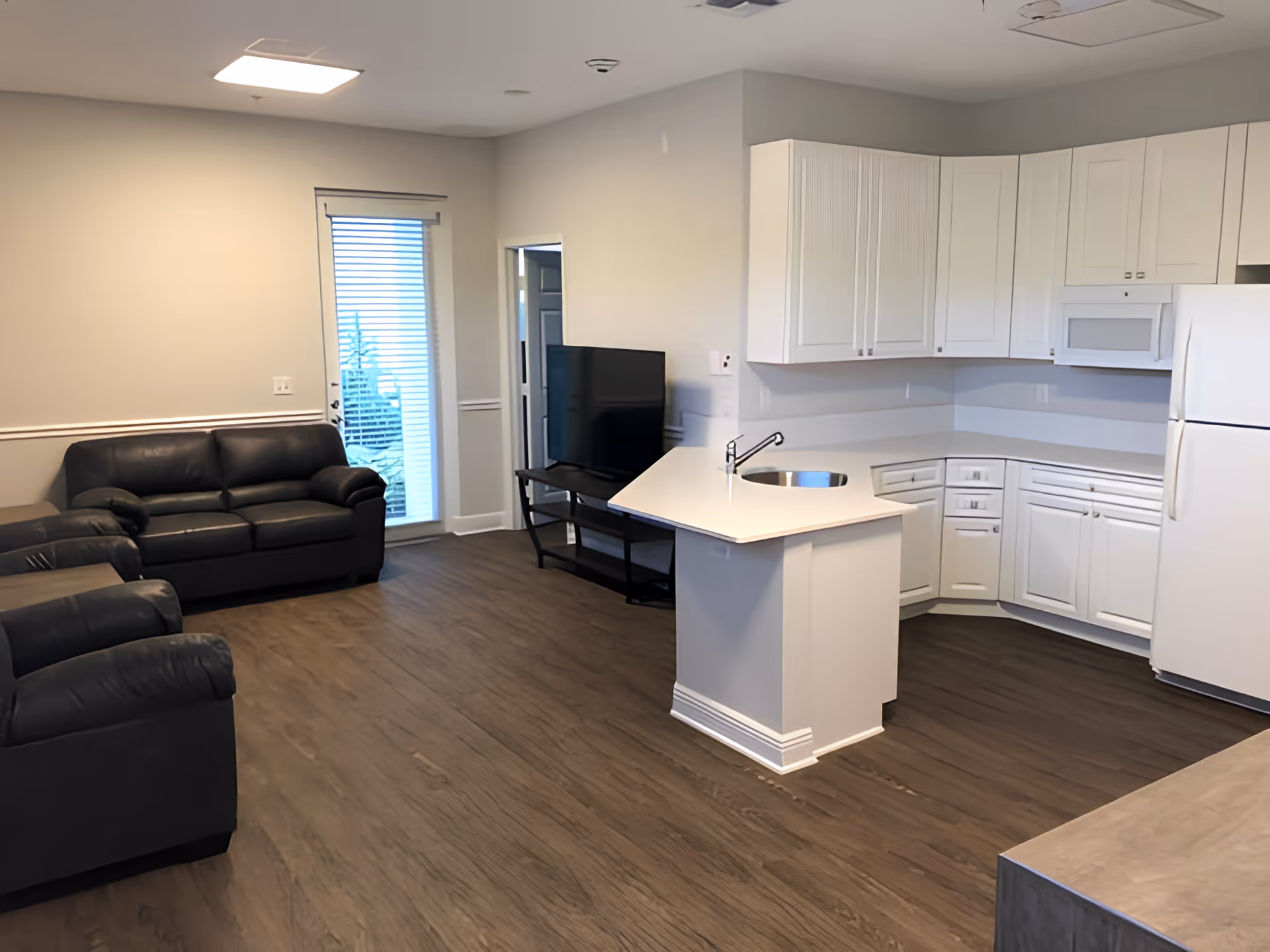 A modern living area with dark leather sofas, a flat-screen TV on a stand, and an open kitchen featuring white cabinets, a white refrigerator, a microwave, and a small island with a sink. The room has wood flooring and a door with blinds leading outside.