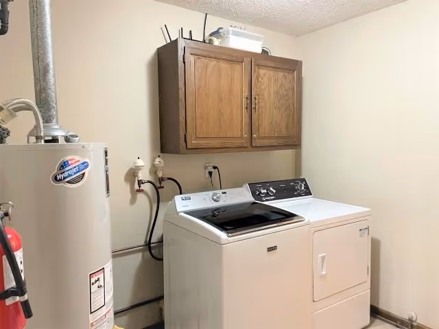 Laundry room with a white washing machine and dryer side by side, a wooden cabinet mounted on the wall above them, and a water heater to the left.