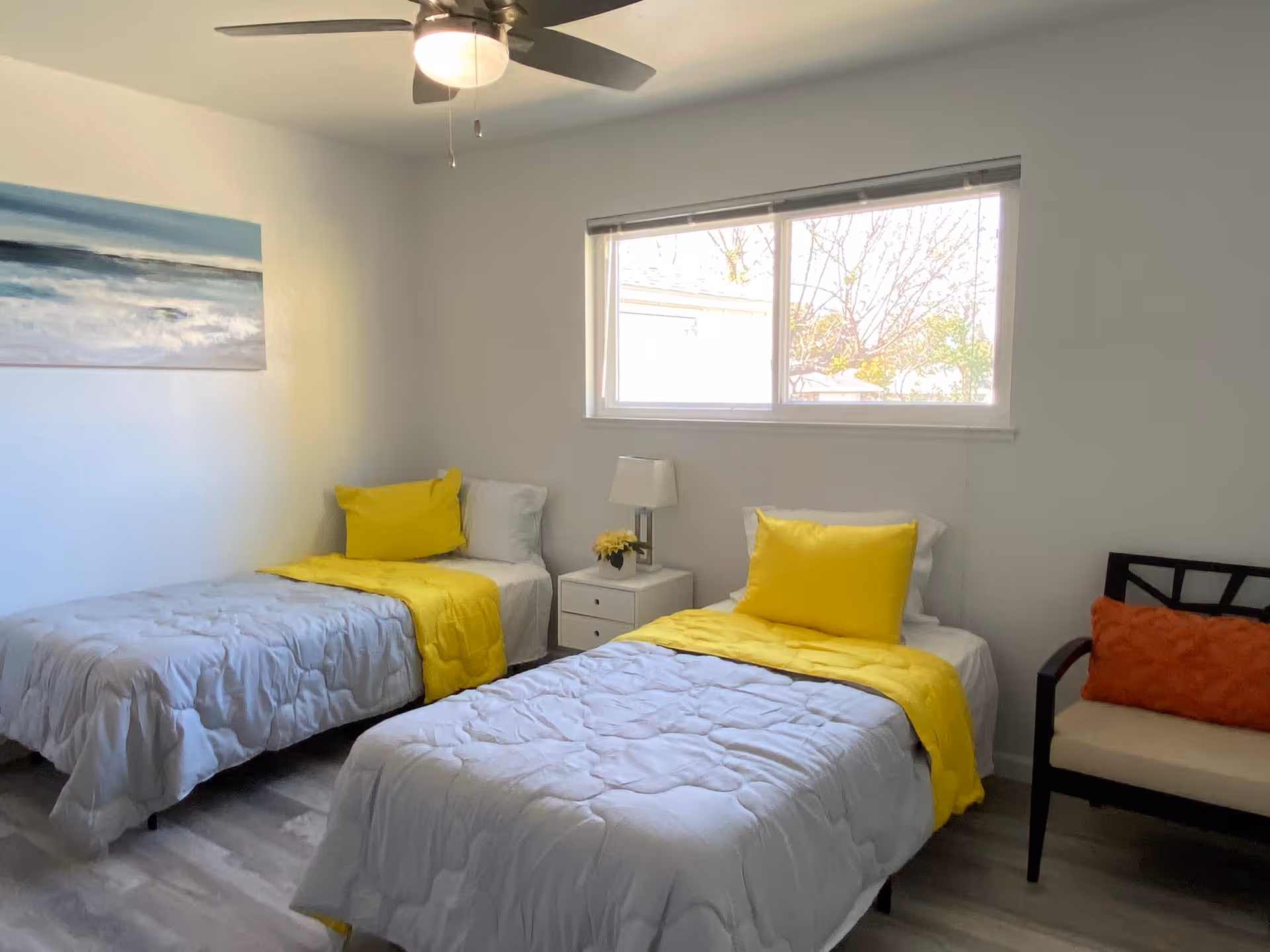 A bright bedroom with two single beds, each made with white bedding and yellow pillows and throws. Between the beds is a small white nightstand with a lamp and a small plant. A window above the nightstand lets in natural light. There is a ceiling fan with a light fixture above, a painting of an ocean scene on the left wall, and a chair with an orange cushion on the right side of the room.