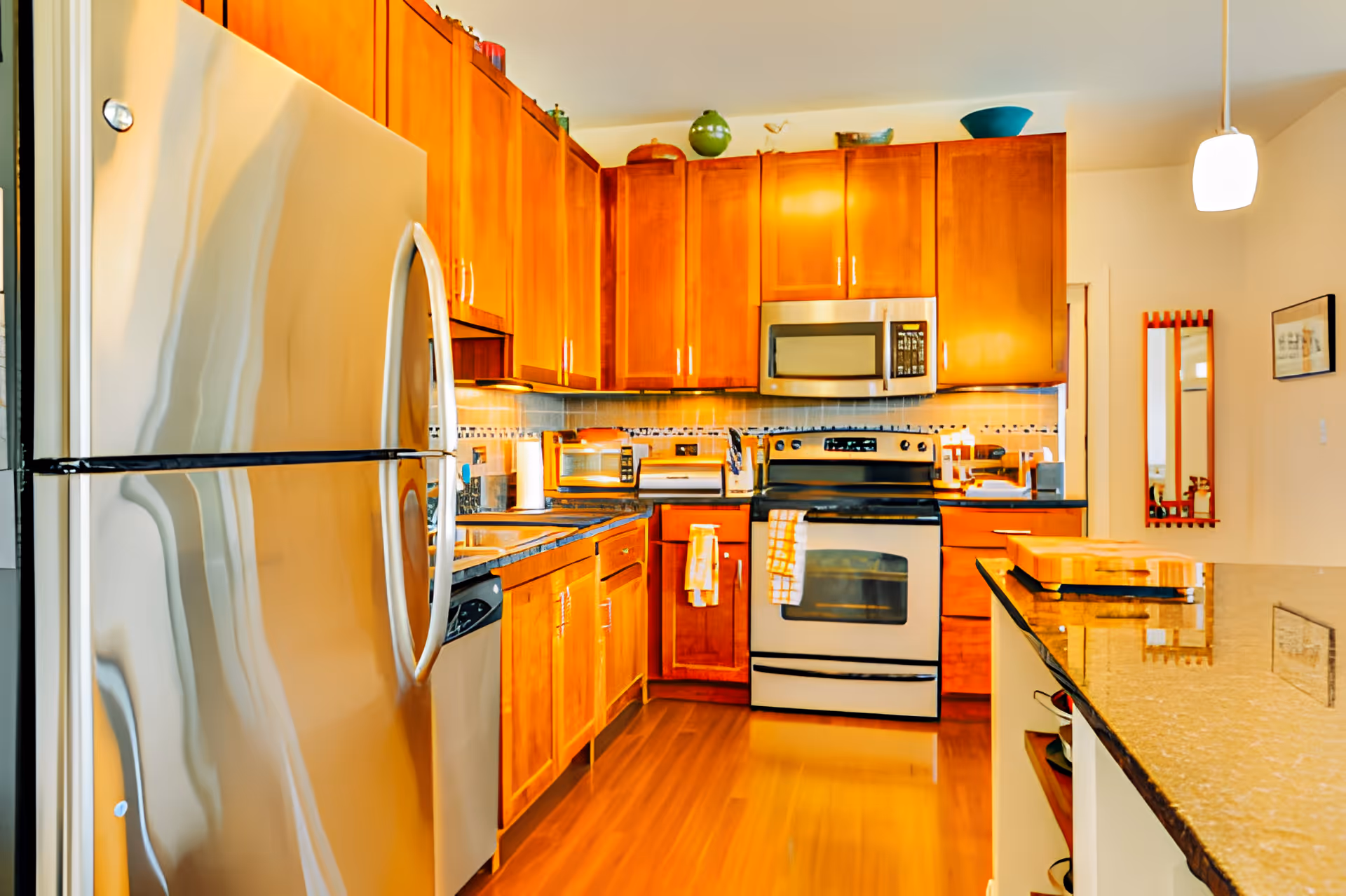 A modern kitchen with wooden cabinets, stainless steel refrigerator, oven, microwave, and dishwasher. The kitchen features a granite countertop island, hardwood flooring, and various kitchen appliances on the counters. A pendant light hangs from the ceiling and a mirror is mounted on the wall in the background.