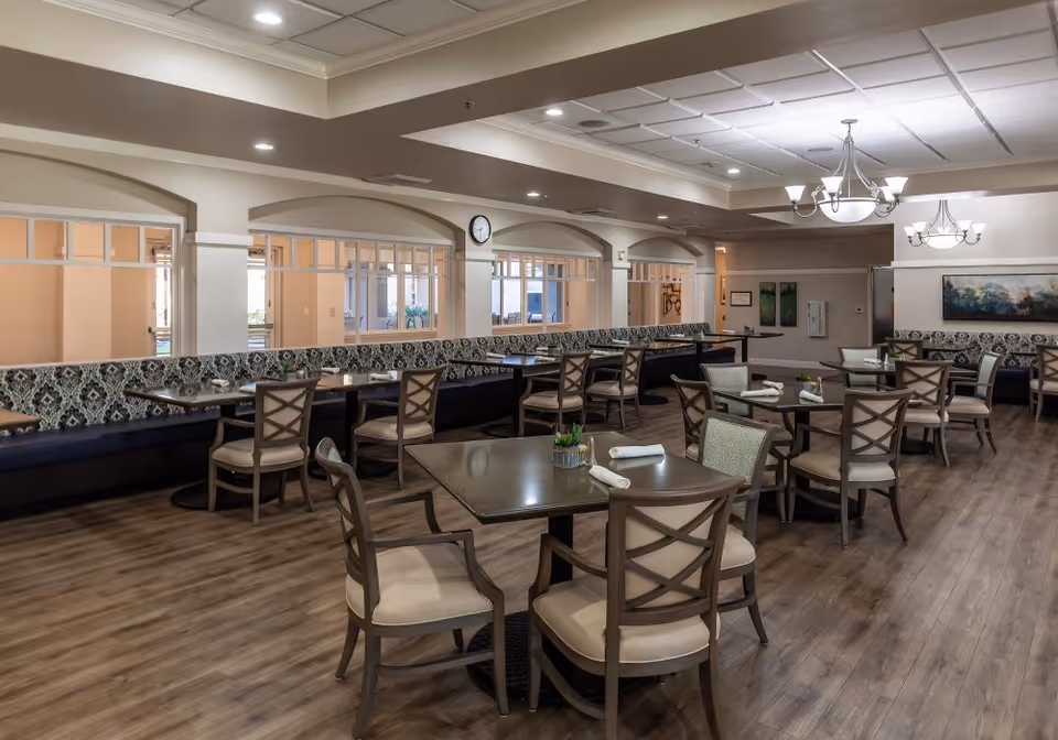 A spacious dining room with multiple tables and chairs arranged neatly. The tables are set with napkins and small decorative plants. The room features patterned bench seating along the wall, wood flooring, and soft overhead lighting with chandeliers. The walls have large windows and artwork, creating a welcoming atmosphere.