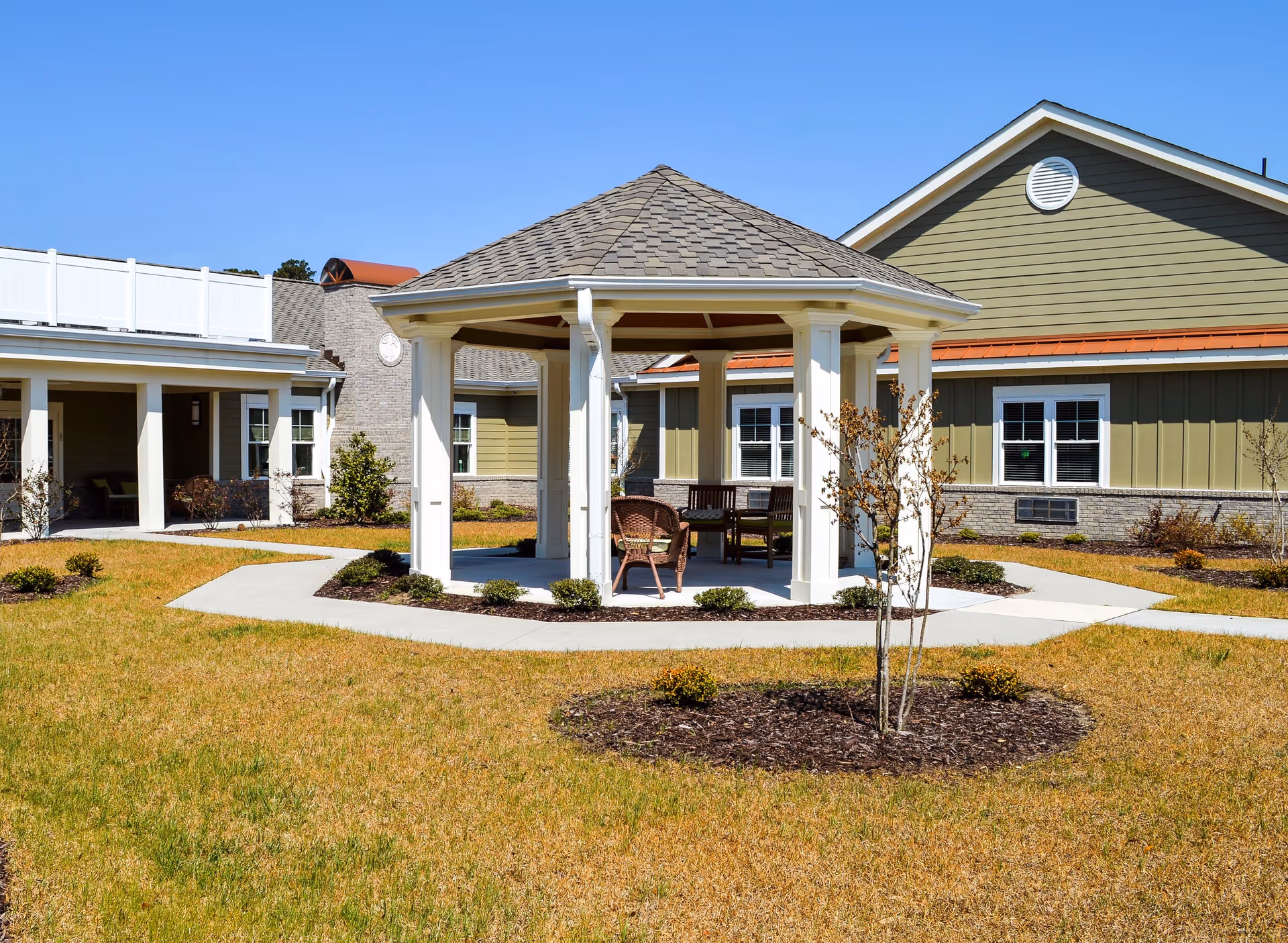 Outdoor courtyard area of a senior living facility featuring a white gazebo with a shingled roof, surrounded by a paved walkway and landscaped with small bushes and young trees. The background shows a single-story building with green siding, white trim, and multiple windows under a clear blue sky.