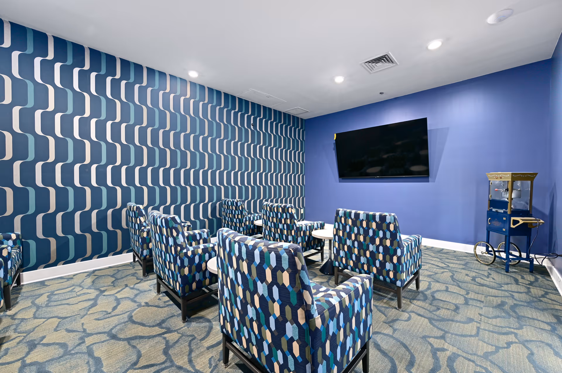A small media room with six patterned armchairs arranged in two rows facing a wall-mounted flat screen TV. The room features a blue accent wall and a patterned wallpaper on one side. There is a blue popcorn machine on a stand in the corner and a carpet with an abstract design covering the floor.