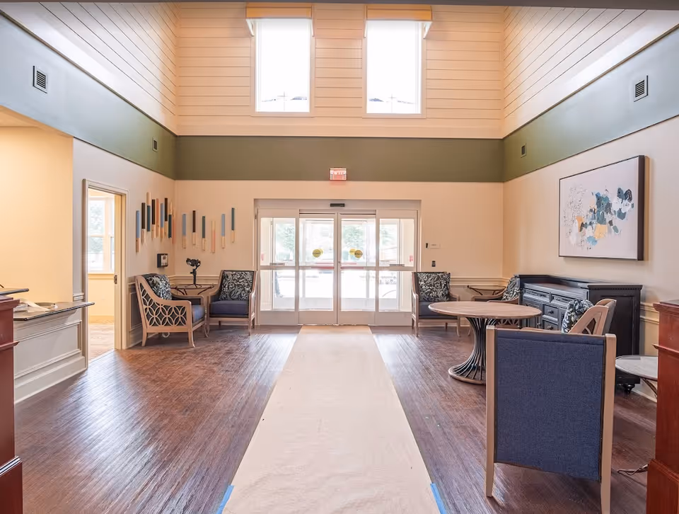A bright and spacious lobby area with high ceilings and two large windows near the ceiling. The room features a wooden floor with a light-colored runner in the center. There are several chairs with patterned cushions arranged around small tables on both sides of the room. A decorative wall art piece is hung on the right wall, and a set of double glass doors leads outside at the far end of the room.