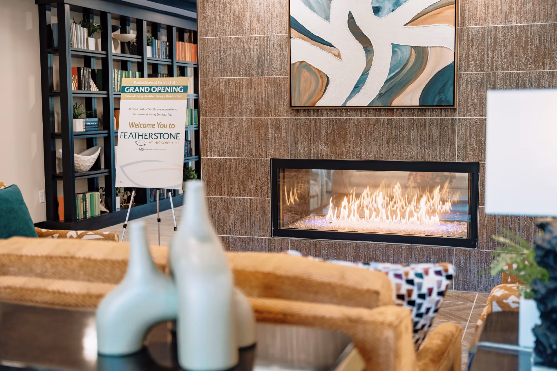 Cozy lounge with a modern gas fireplace, abstract painting above, seating in the foreground and a 'Grand Opening' sign for FeatherStone at Hickory Hill.
