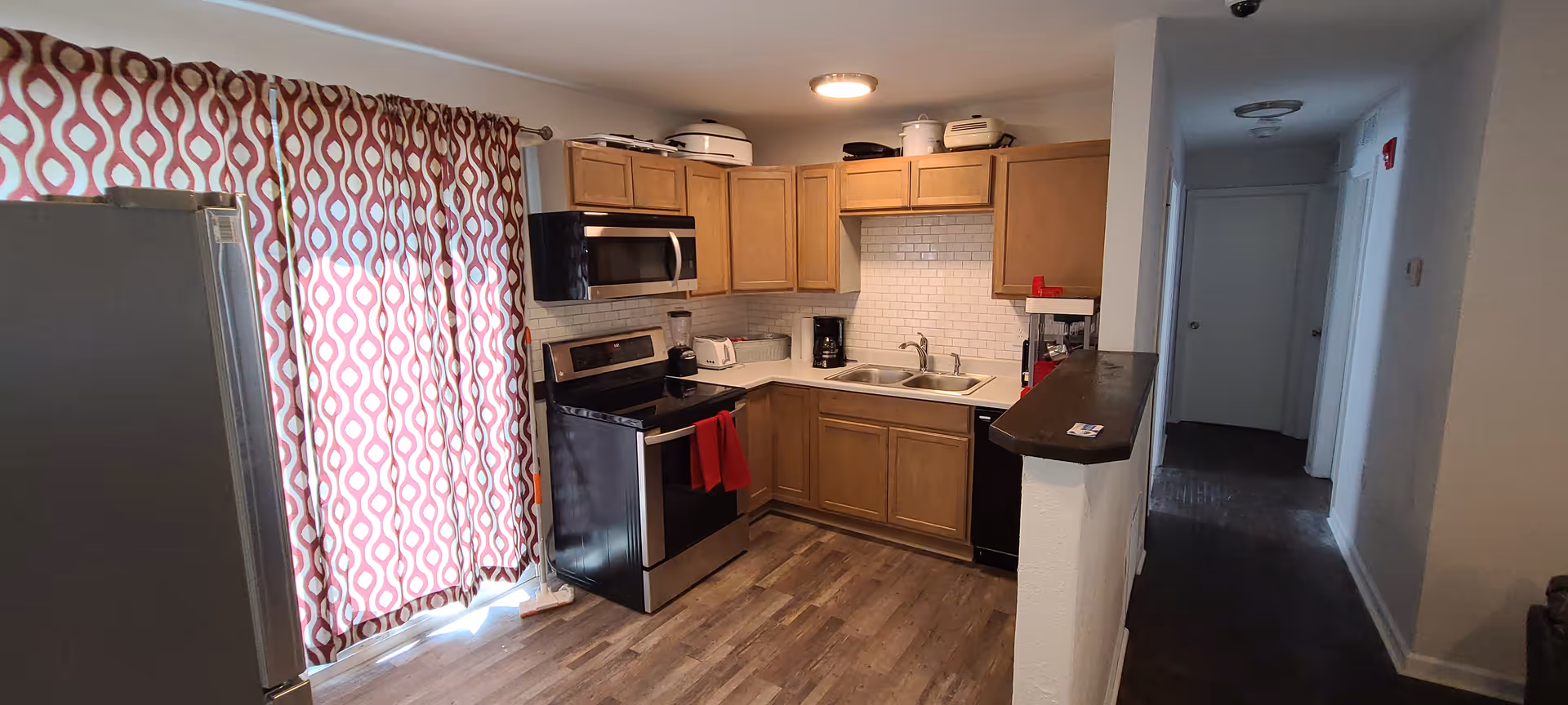 Open kitchen with wooden cabinets, stainless steel appliances, double sink and red patterned curtains over a sliding door.