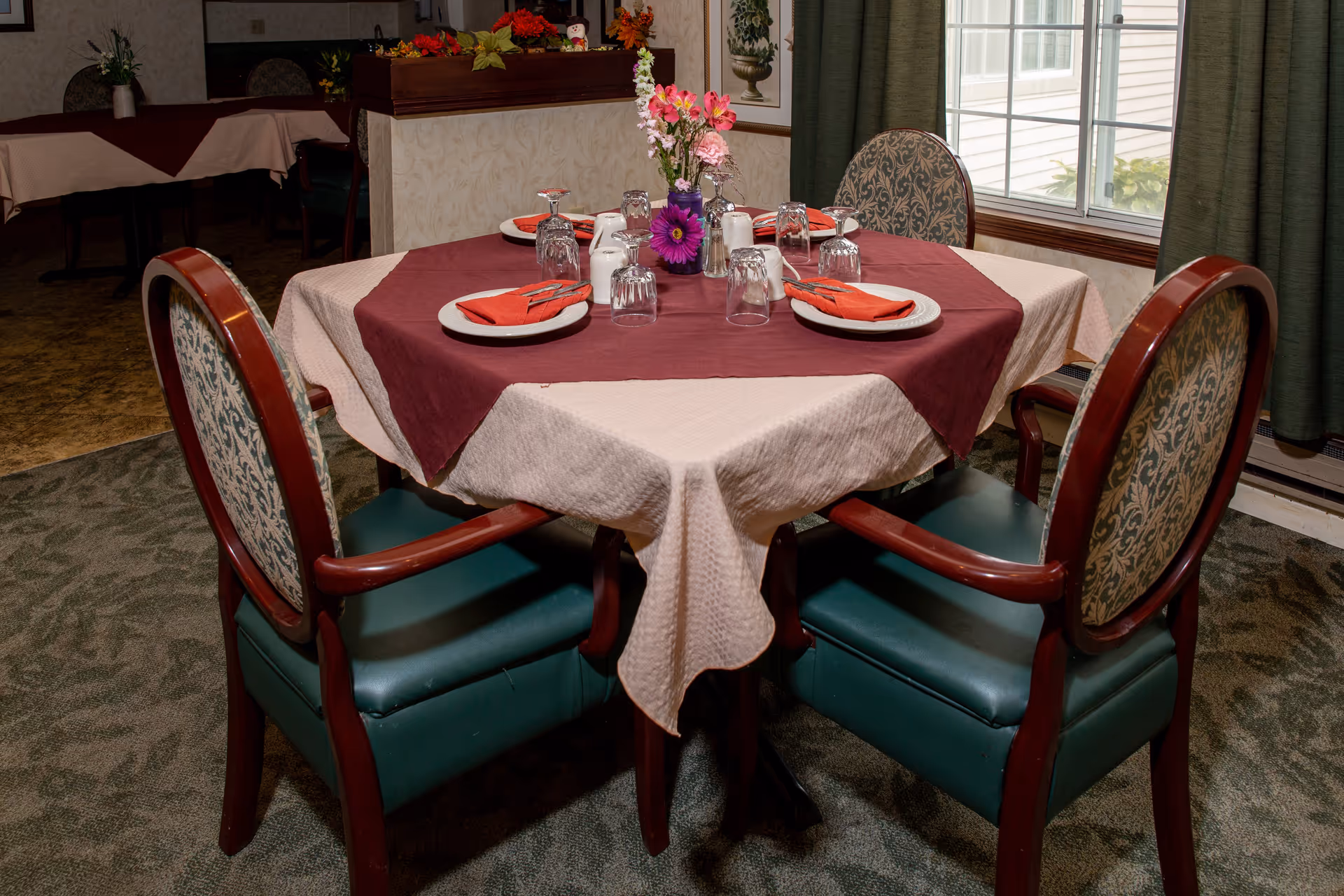 Round dining table set for four with burgundy and beige tablecloths, plates, glasses, napkins, and a floral centerpiece in a dining area.