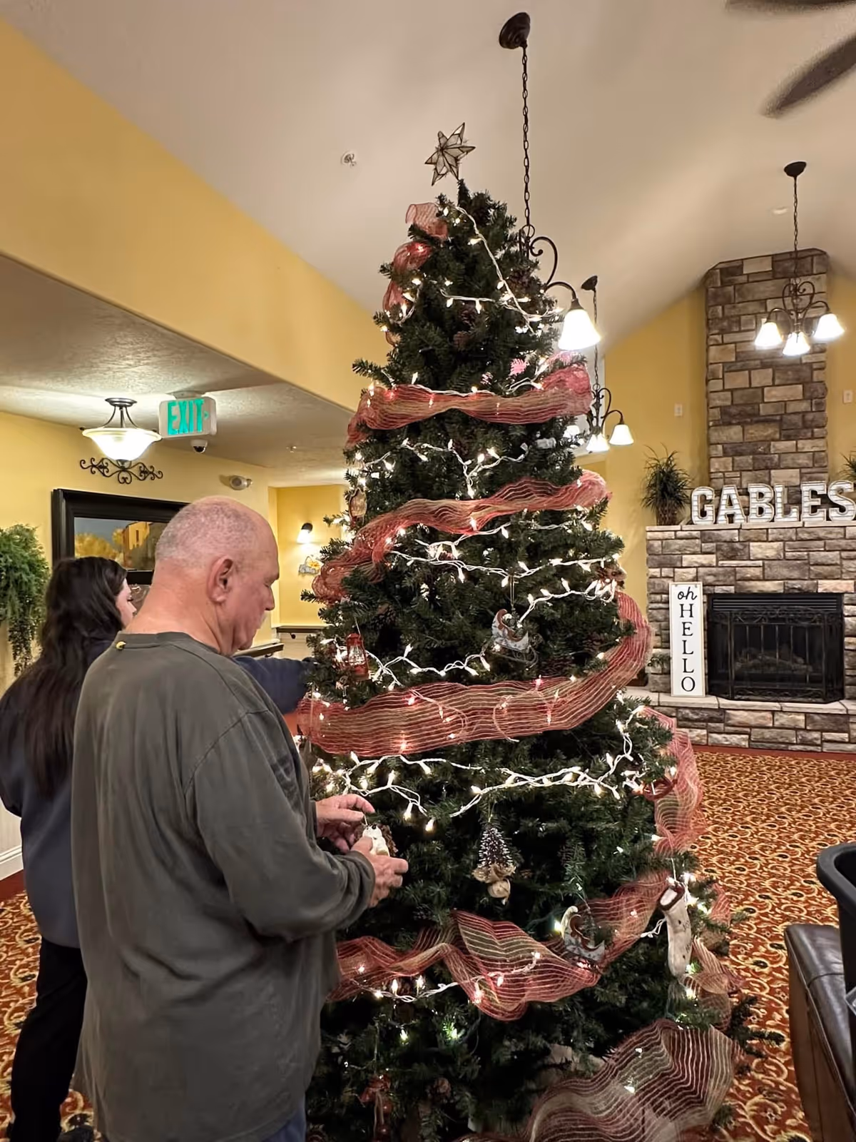 Two people decorating a large Christmas tree with white lights and red ribbon in a warmly lit room with yellow walls. The room features a stone fireplace with the word 'GABLES' displayed on the mantel and a sign next to it that says 'oh HELLO'.