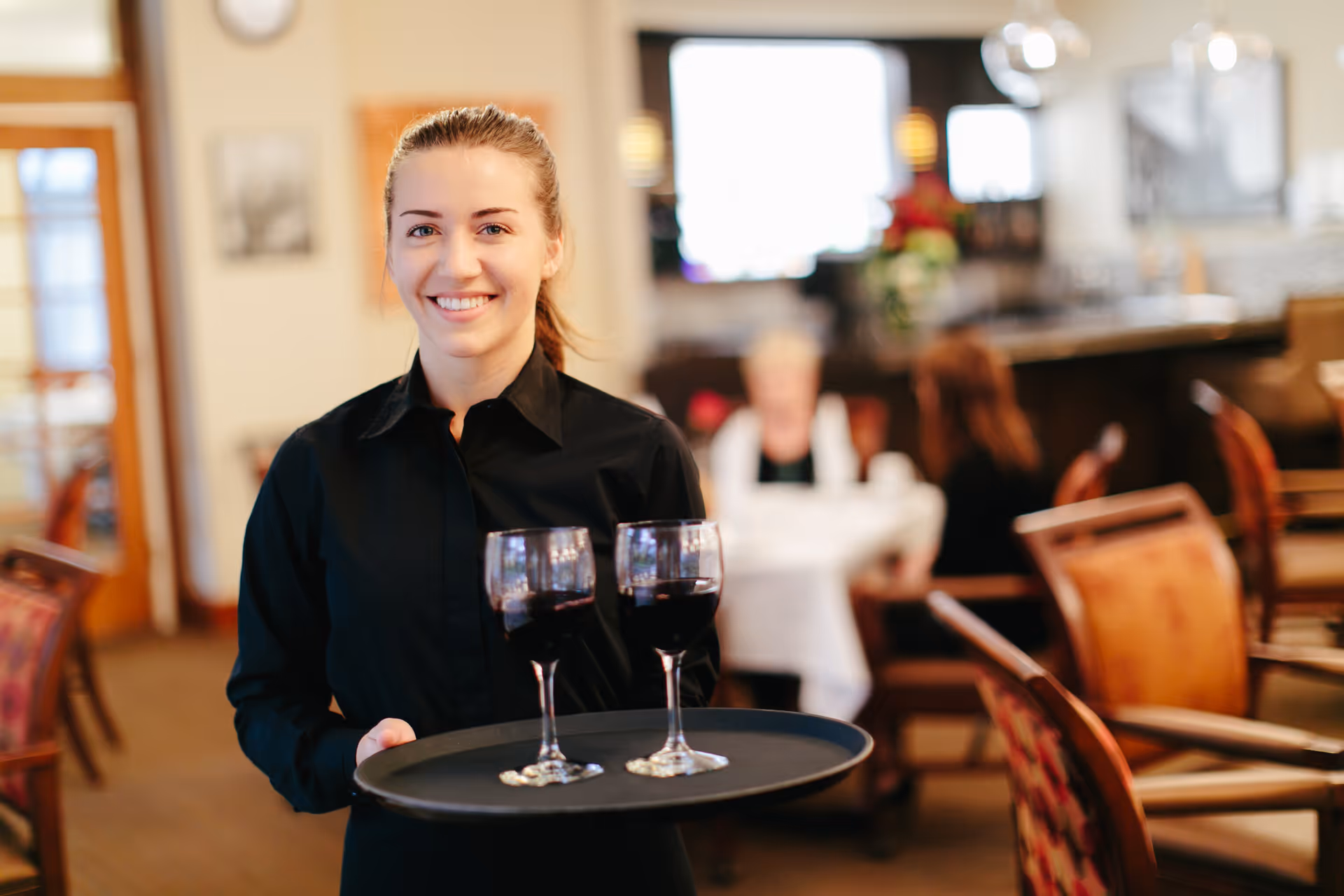 A smiling server in a black shirt holding a tray with two glasses of red wine in a dining room with tables and chairs.
