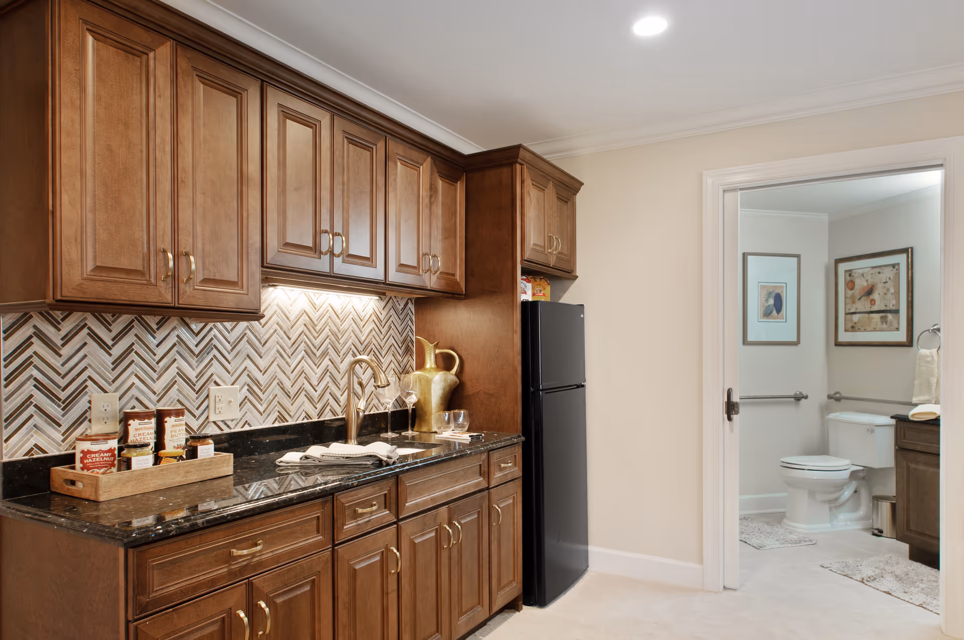 A kitchen area with wooden cabinets, a black granite countertop, a sink with a gold faucet, and a black refrigerator. The backsplash features a herringbone tile pattern. To the right, there is an open doorway leading to a bathroom with a toilet, framed artwork on the walls, and a towel hanging on a ring.
