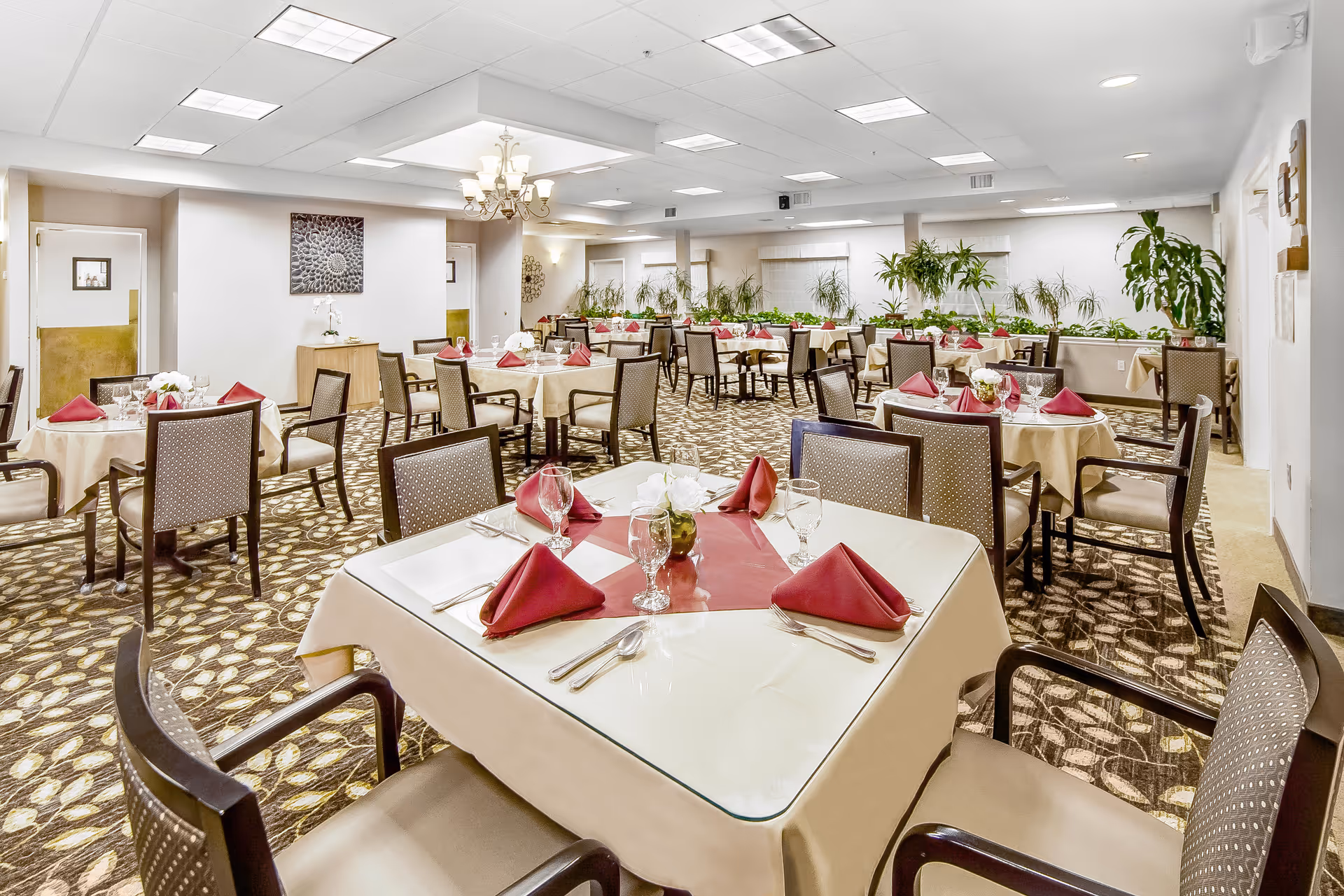 A spacious dining room in a senior living facility with multiple tables covered in beige tablecloths and set with red folded napkins, glassware, and silverware. The room features patterned carpet, several chairs around each table, potted plants along the walls, and a chandelier hanging from the ceiling.