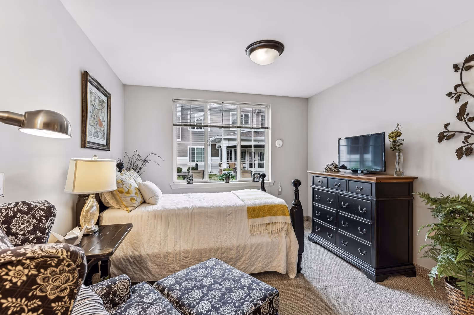 Bright furnished bedroom with a bed under a window, a patterned armchair and ottoman, and a dark dresser topped with a TV.