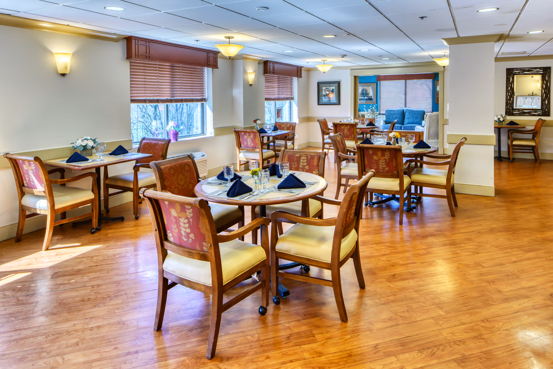 A bright dining room with wooden floors and multiple round and square tables set with glasses, napkins, and silverware. The chairs have wooden frames with cushioned seats and backs featuring a floral pattern. Windows with wooden blinds allow natural light to fill the room. Wall sconces and ceiling lights provide additional lighting. In the background, a seating area with blue upholstered furniture is visible.