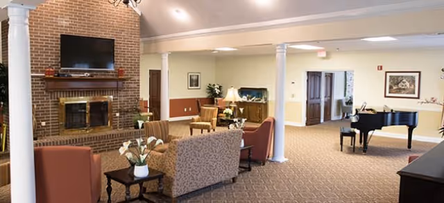 A cozy lobby area featuring comfortable seating, a fireplace with a television above it, and a piano. The space is well-lit with decorative plants and a warm color palette.